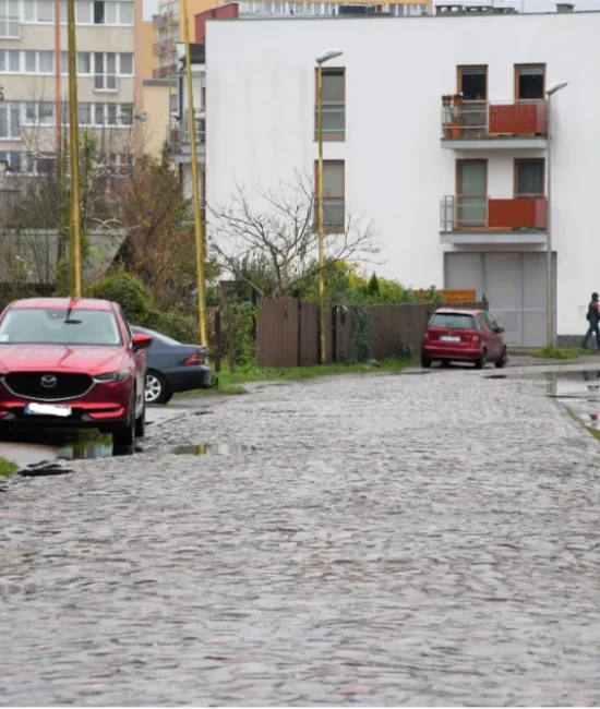 Niska, brukowana ulica między budynkami mieszkalnymi, z zaparkowanymi samochodami po obu stronach.