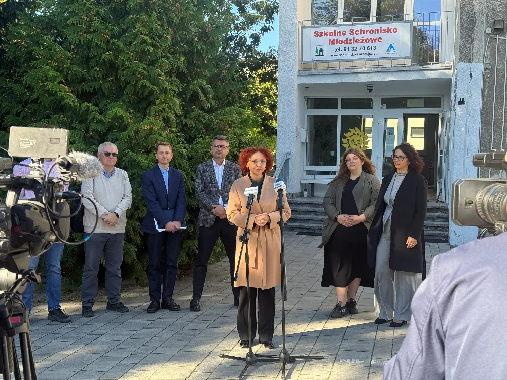 Kilka osób stoi przed wejściem do budynku schroniska młodzieżowego podczas konferencji prasowej.