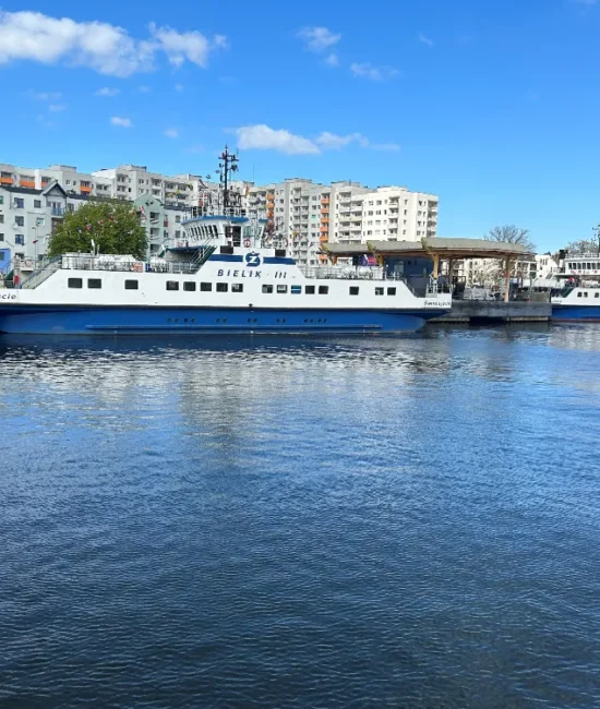 Prom Bielik III zacumowany przy nabrzeżu nad wodą, w tle zabudowa miejska i drugi prom.