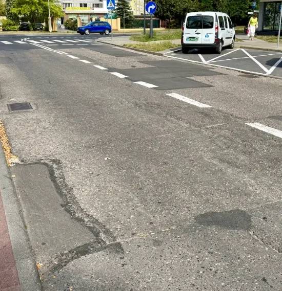 Zniszczona jezdnia uliczna z oznakowaniem poziomym i zaparkowanymi samochodami.