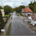 Widok wąskiej ulicy z zabudową jednorodzinną i jezdnią po deszczu.