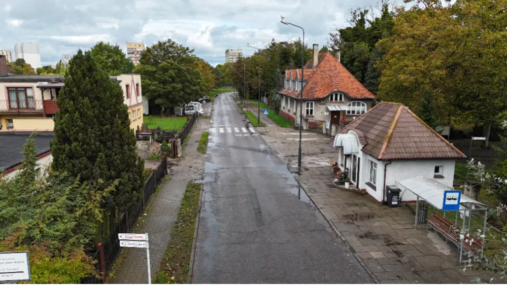 Widok wąskiej ulicy z zabudową jednorodzinną i jezdnią po deszczu.