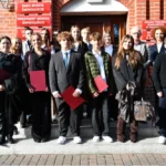 Grupa uczniów stoi przed wejściem do budynku urzędu miejskiego z czerwonymi teczkami w rękach.